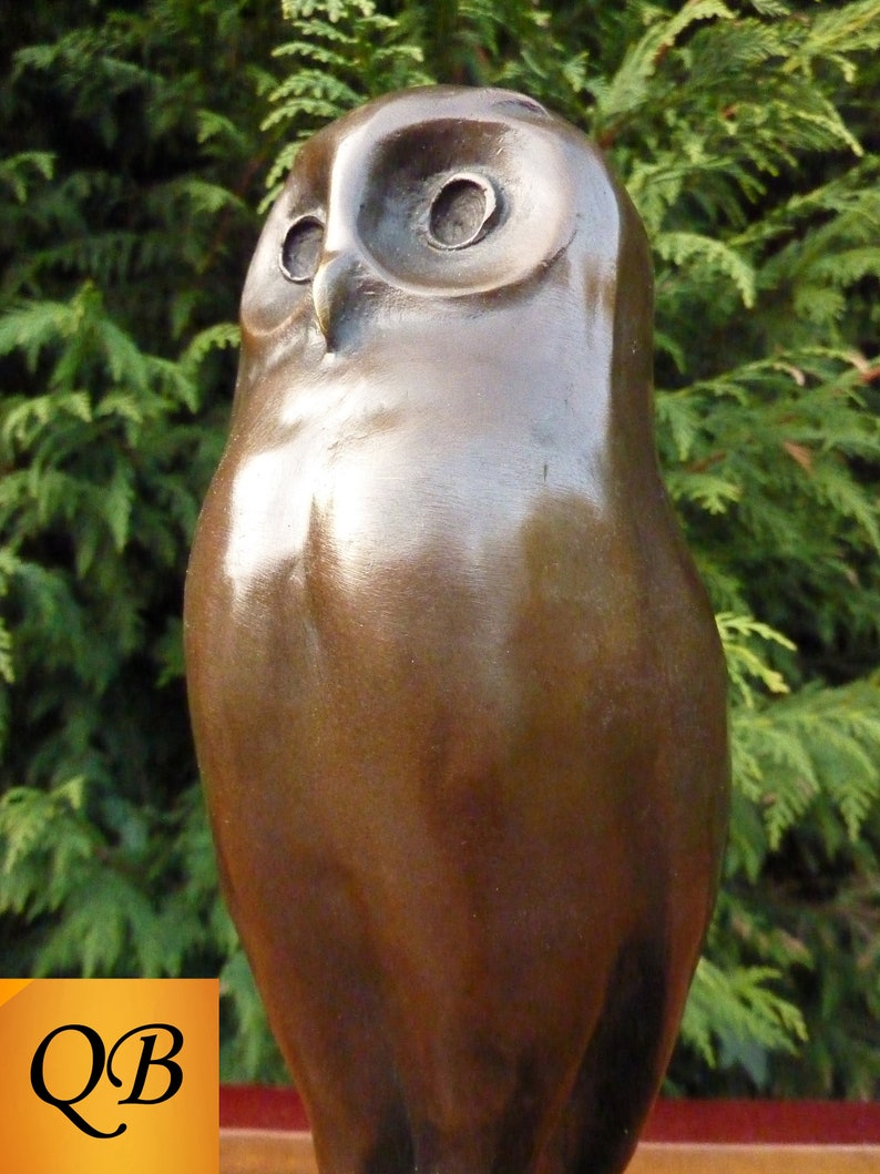 May include: A bronze-colored owl sculpture with a smooth, polished finish. The owl has large, round eyes and a slightly tilted head. The background is a blurred green hedge. The letters "QB" are in the lower left corner.