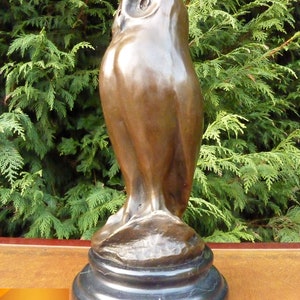 May include: A bronze owl sculpture with a smooth, polished finish. The owl is perched on a black, circular base. The sculpture has a detailed depiction of the owl's features, including its eyes and feathers. The background is a green hedge.