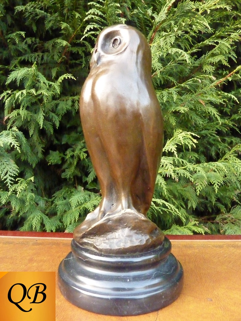 May include: A bronze owl sculpture with a smooth, polished finish. The owl is perched on a black, circular base. The sculpture has a detailed depiction of the owl's features, including its eyes and feathers. The background is a green hedge.