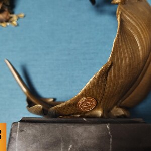 May include: A bronze-colored sculpture of a feather, mounted on a dark gray marble base. The feather has detailed veining and a small, circular emblem. The sculpture is set against a blue background.