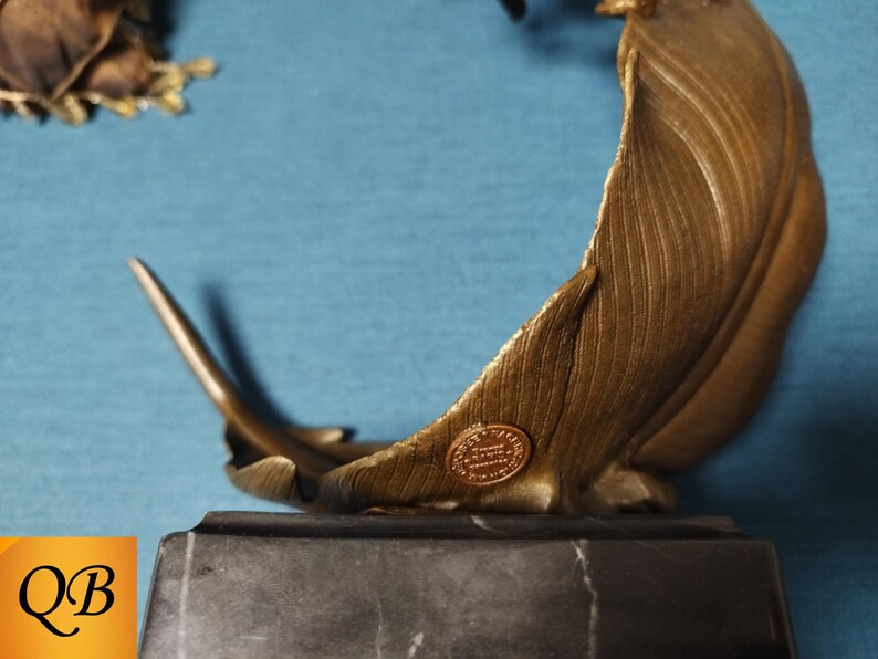May include: A bronze-colored sculpture of a feather, mounted on a dark gray marble base. The feather has detailed veining and a small, circular emblem. The sculpture is set against a blue background.