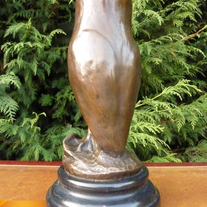 May include: A bronze owl sculpture with a smooth, polished finish. The owl is perched on a black marble base. The sculpture has a classic, elegant design, suitable for home decor. The background features green foliage.