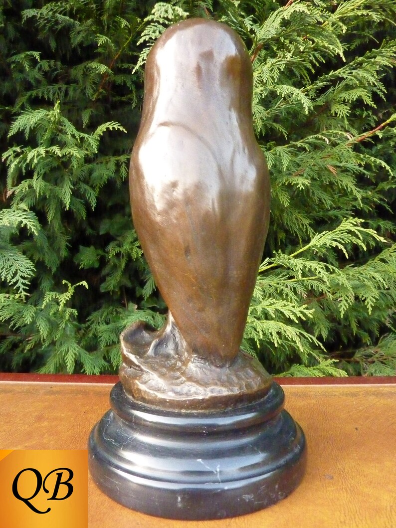 May include: A bronze owl sculpture with a smooth, polished finish. The owl is perched on a black marble base. The sculpture has a classic, elegant design, suitable for home decor. The background features green foliage.
