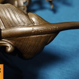 May include: Close-up of a bronze-colored sculpture featuring a detailed feather. The feather has intricate veining and the word "Ancomette" is inscribed on the surface. The sculpture is set against a blue background.