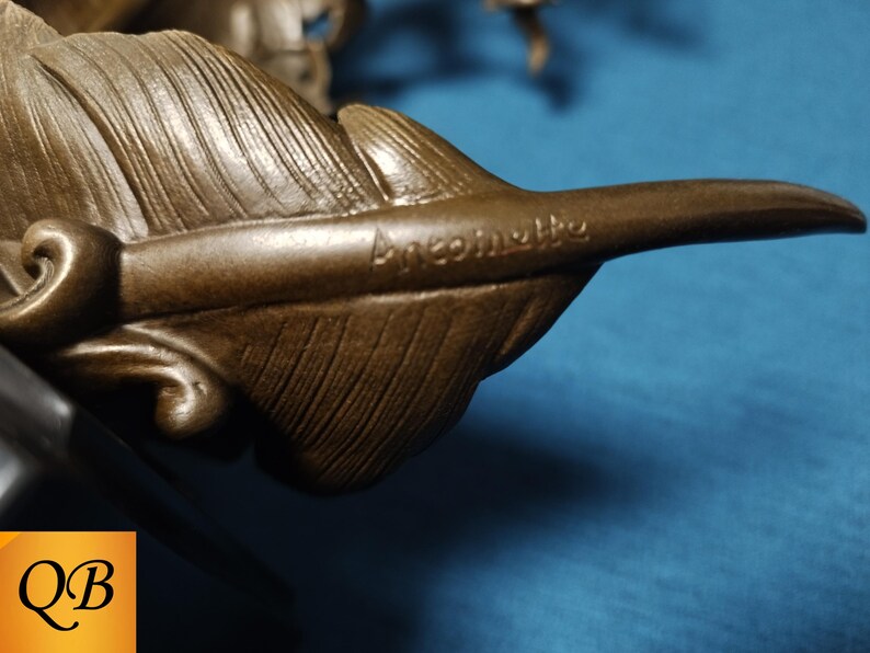 May include: Close-up of a bronze-colored sculpture featuring a detailed feather. The feather has intricate veining and the word "Ancomette" is inscribed on the surface. The sculpture is set against a blue background.