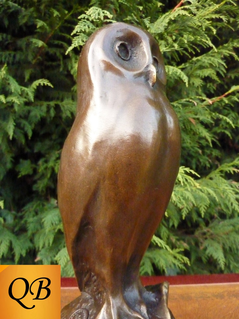 May include: A bronze owl sculpture with a smooth, reflective surface. The owl is depicted in a standing pose, with detailed eyes and a slightly forward-facing gaze. The sculpture is set against a backdrop of green foliage.