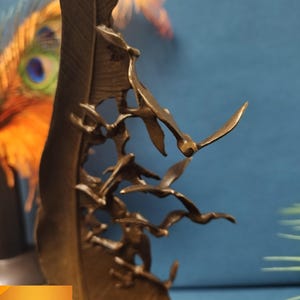 May include: Bronze sculpture of a large feather with a flock of birds in flight. The birds are intricately detailed, with outstretched wings. The sculpture has a warm, metallic finish and is set against a blue background.