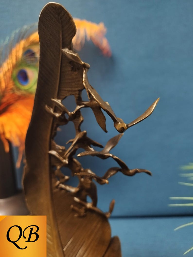 May include: Bronze sculpture of a large feather with a flock of birds in flight. The birds are intricately detailed, with outstretched wings. The sculpture has a warm, metallic finish and is set against a blue background.