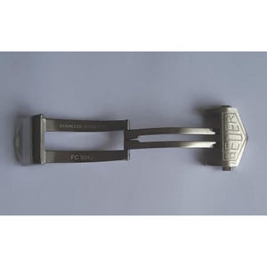 May include: A silver watch band clasp with the word "HEUER" engraved on the side. The clasp has two prongs and the text "STAINLESS STEEL FC 5042" is engraved on the bottom.