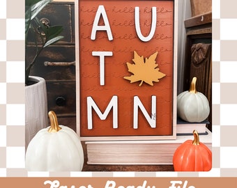 May include: A wooden sign with the word "AUTUMN" in white letters with a brown background. The letters are raised and have a 3D effect. There is a small orange maple leaf in the centre of the sign. The sign is sitting on a stack of books. The background is a checkered pattern.