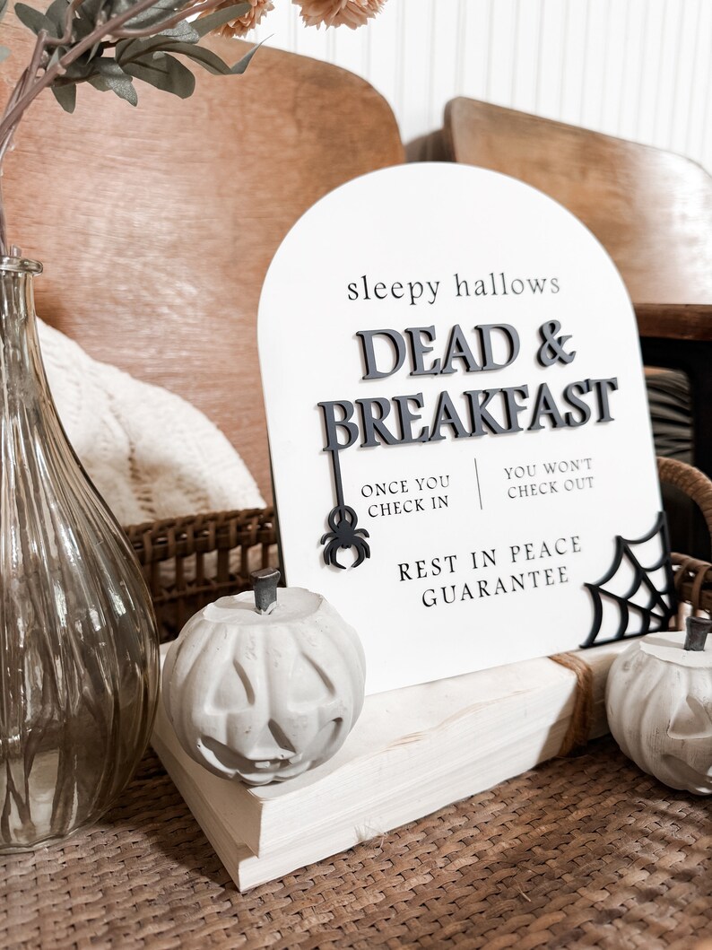 May include: A white sign with black text that reads "sleepy hallows DEAD & BREAKFAST" with a spider and web graphic. The sign is propped up on a stack of white books with two concrete pumpkin decorations.