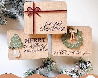 May include: Three wooden Christmas signs with laser-cut designs. The first sign says "merry christmas" with a red ribbon bow. The second sign says "MERRY everything & happy always" with a green Christmas tree and a gift box. The third sign says "a little gift for you" with a green wreath and a red bow.