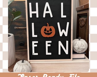 May include: A black sign with the word "HALLOWEEN" in white letters. A small orange pumpkin with a carved face is in the centre of the sign. The sign is framed in wood and is sitting on a stack of books.