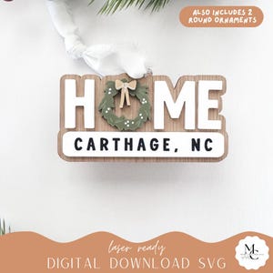 Puede incluir: Un letrero de madera HOME con letras blancas y una corona verde con un lazo. El letrero incluye el texto "CARTHAGE, NC". La imagen también incluye el texto "ALSO INCLUDES 2 ROUND ORNAMENTS" y "laser ready DIGITAL DOWNLOAD SVG".