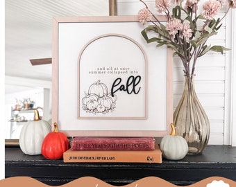 May include: A laser-cut wood sign with a fall theme. The sign features a black arch with the text "and all at once summer collapsed into fall" and a black outline of pumpkins. The sign is sitting on a stack of books on a black surface.
