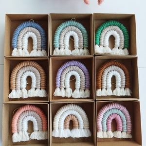 May include: Nine macrame rainbow keyrings in individual cardboard boxes. Each keyring features a different colour scheme, including blue, green, brown, and pink. The keyrings have a looped top for hanging and a fringe at the bottom.