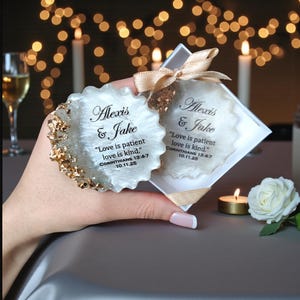 May include: Two wedding favours, one round and one square, both with the names "Alexis & Jake" and the quote "Love is patient love is kind." printed on them. The round coaster has gold accents. The square one is in a box tied with a ribbon.
