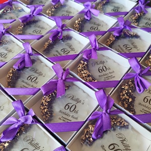 May include: A collection of square white gift boxes, each containing a round white resin coaster with gold-colored metallic edges and purple ribbon bows. The coasters are printed with the text Phillip's 60th Birthday Bash, Zanzibar, 10/30/2025.