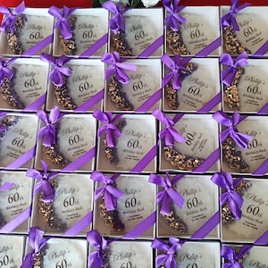 May include: A collection of square white gift boxes, each containing a circular white resin coaster with gold and purple accents. Each coaster features a purple ribbon bow and printed text reading Phillip's 60th Birthday Bash, Zanzibar, 10/30/2025.