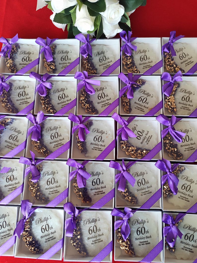 May include: A collection of square white gift boxes, each containing a circular white resin coaster with gold and purple accents. Each coaster features a purple ribbon bow and printed text reading Phillip's 60th Birthday Bash, Zanzibar, 10/30/2025.