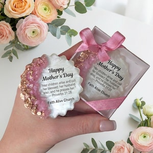 Könnte beinhalten: Ein Muttertags-Geschenkset mit einem dekorativen, runden Gegenstand mit dem Text "Happy Mother's Day" und einem Bibelvers. Der Artikel hat rosa und goldfarbene Akzente. Eine transparente Box mit rosa Schleife und einem ähnlichen Artikel ist ebenfalls zu sehen, umgeben von rosa und pfirsichfarbenen Rosen.