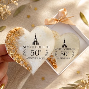 May include: Two heart-shaped resin ornaments with gold leaf accents, each printed with a church icon and the text NORTH CHURCH 50th ANNIVERSARY. One ornament is held up while the other rests inside a white gift box with a light gold ribbon bow.