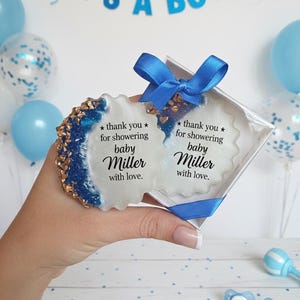 Op de afbeelding: Een babyshower cadeau set met twee witte en blauwe onderzetters in een doorzichtige doos, vastgebonden met een blauw lint. De onderzetters hebben gouden en blauwe accenten en de tekst "bedankt voor het showeren van baby Miller met liefde."
