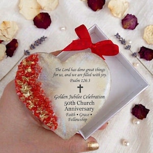 May include: A heart-shaped keepsake with red and gold accents, presented in a white box with a red ribbon. The keepsake has text that reads "The Lord has done great things for us, and we are filled with joy. Psalm 126:3" and "Golden Jubilee Celebration 50th Church Anniversary Faith • Grace • Fellowship."