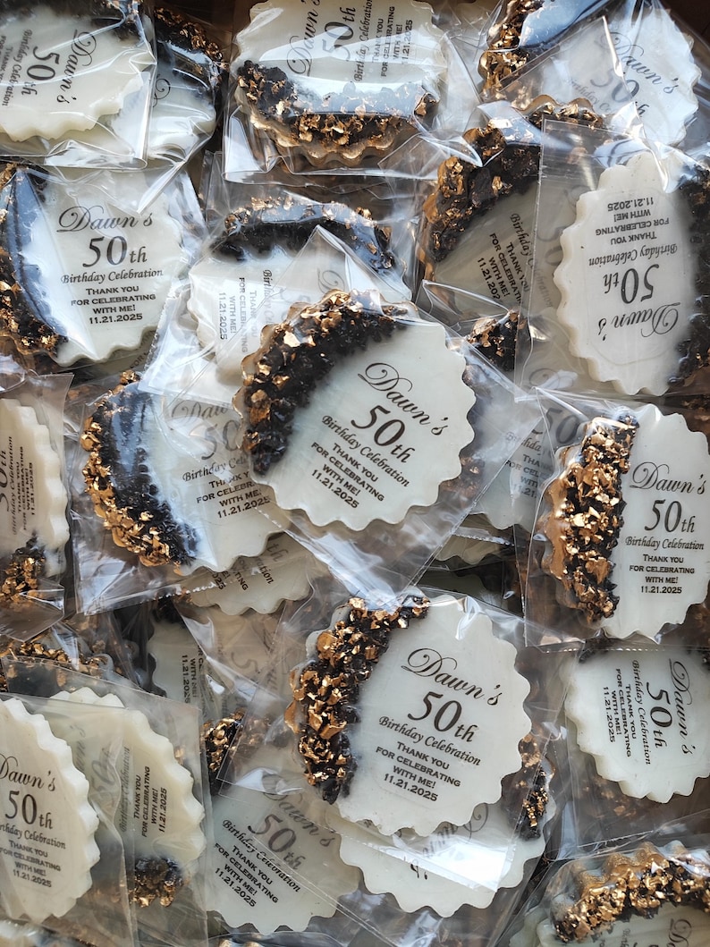 May include: A collection of round, scalloped-edge white party favors individually wrapped in clear plastic. Each favor features black text reading Dawn's 50th Birthday Celebration, Thank You For Celebrating With Me, 11.21.2025, and a crescent-shaped accent of black and gold metallic flakes.