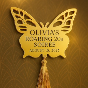 May include: A gold-colored butterfly-shaped sign with the text "OLIVIA'S ROARING 20s SOIRÉE AUGUST 15, 2025". The sign has a decorative tassel hanging from the bottom. The background is a patterned, dark gold color.