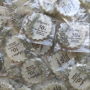 May include: A collection of round, scalloped-edge white resin coasters individually wrapped in clear plastic. Each coaster features silver metallic leaf accents on one side and black printed text reading Sophia's 50th Birthday Celebration THANK YOU FOR COMING.