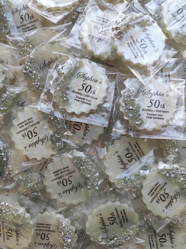 May include: A collection of round, scalloped-edge white resin coasters individually wrapped in clear plastic. Each coaster features silver metallic leaf accents on one side and black printed text reading Sophia's 50th Birthday Celebration THANK YOU FOR COMING.
