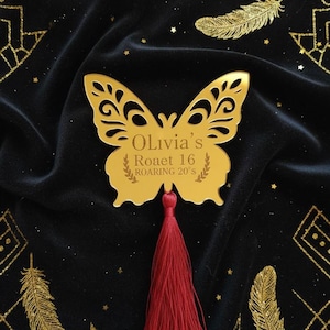 May include: A gold butterfly-shaped decoration with intricate cut-out details, featuring the text "Olivia's Roaet 16 ROARING 20's". It has a red tassel and is set against a black background with gold feather and geometric designs.