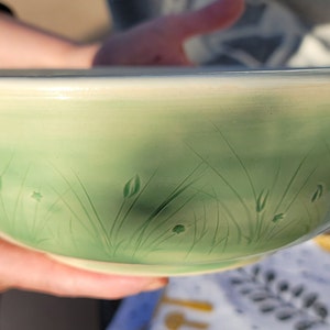 May include: A green ceramic bowl with a subtle ombre effect, featuring a hand-painted design of grass and flowers.
