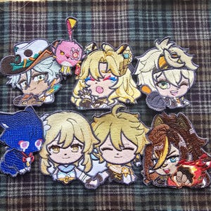 May include: A collection of embroidered patches depicting anime-style characters. The patches feature detailed facial expressions and colourful outfits. The designs include characters with blonde and brown hair, and a blue cat-like figure. The patches are displayed on a plaid background.