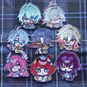 May include: A collection of embroidered patches depicting anime-style characters. The patches feature detailed facial expressions and vibrant hair colours, including blue, grey, red, and purple. Each character has a unique outfit. The patches are displayed on a blue and black plaid fabric.