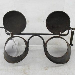 May include: An antique pair of spectacles with a dark, aged metal frame. The design includes two large, circular lenses, two circular shades, and two cone-shaped ear pieces. The frame has a unique, vintage aesthetic.