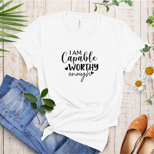 May include: White t-shirt with black text that reads "I am Capable Worthy Enough" with two small hearts on either side of the word "worthy".
