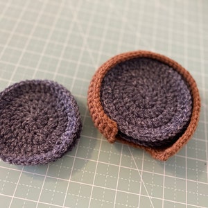 May include: Two crocheted coasters, one with a brown border and one without. Both coasters are a dark gray color.