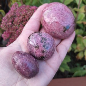 May include: Three polished rhodonite stones of varying sizes, each featuring a deep pink base colour with dark black and brown mineral inclusions. The stones have a smooth, rounded texture and are arranged in a line from smallest to largest.