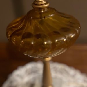 May include: A gold-toned bottle stopper with a round, amber-colored glass top. The top has a textured, rippled design. The stopper is on a white base.