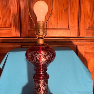 May include: A ruby red glass table lamp with a decorative floral pattern etched into the glass. The lamp has a brass base and a brass neck. The lamp is lit with a standard light bulb.