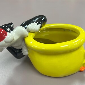 May include: A yellow ceramic planter shaped like a bird with a black cat figurine peeking out of the top. The cat has a white face and red nose.