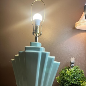 May include: A light blue Art Deco style table lamp with a fan-shaped base. The lamp has a gold-colored neck and a clear, rectangular finial. The bulb is lit, and the background is a warm brown.