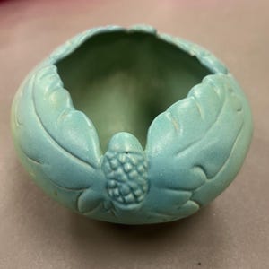 May include: A small, round, turquoise ceramic bowl with a leaf and acorn design. The bowl has a smooth, glossy finish and a shallow interior. The exterior features detailed leaf-shaped carvings and a small acorn at the base.