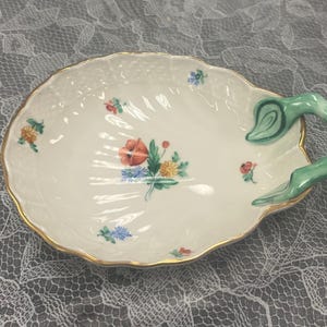 May include: Oval-shaped, off-white ceramic dish with a gold rim and a green handle. The dish is decorated with colorful floral bouquets. The handle is a curved, stylized leaf shape. A decorative serving piece.