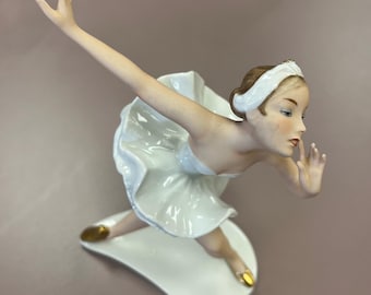 May include: A porcelain figurine of a ballerina in a white tutu and headband. The ballerina is posed mid-dance, with one arm extended and the other near her face. Her ballet slippers are gold. The figurine is on a white base.