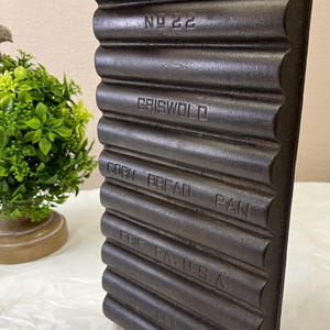 May include: A vintage cast iron corn bread pan with a ribbed design. The pan is dark brown and features the embossed text "NO 22", "GRISWOLD", "CORN BREAD PAN", and "ERIE PA. U.S.A."