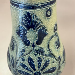 May include: A ceramic vase with a light beige background and dark blue floral patterns. The vase has a narrow neck and a wide body, with a textured surface. The floral design includes flowers and leaves.