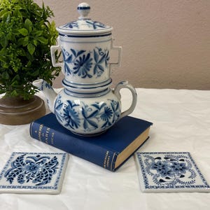 May include: A white and blue porcelain tea set with floral designs, including a teapot, infuser, and lid. Two matching square coasters are also visible. The set is placed on a blue book, with a green plant in the background.